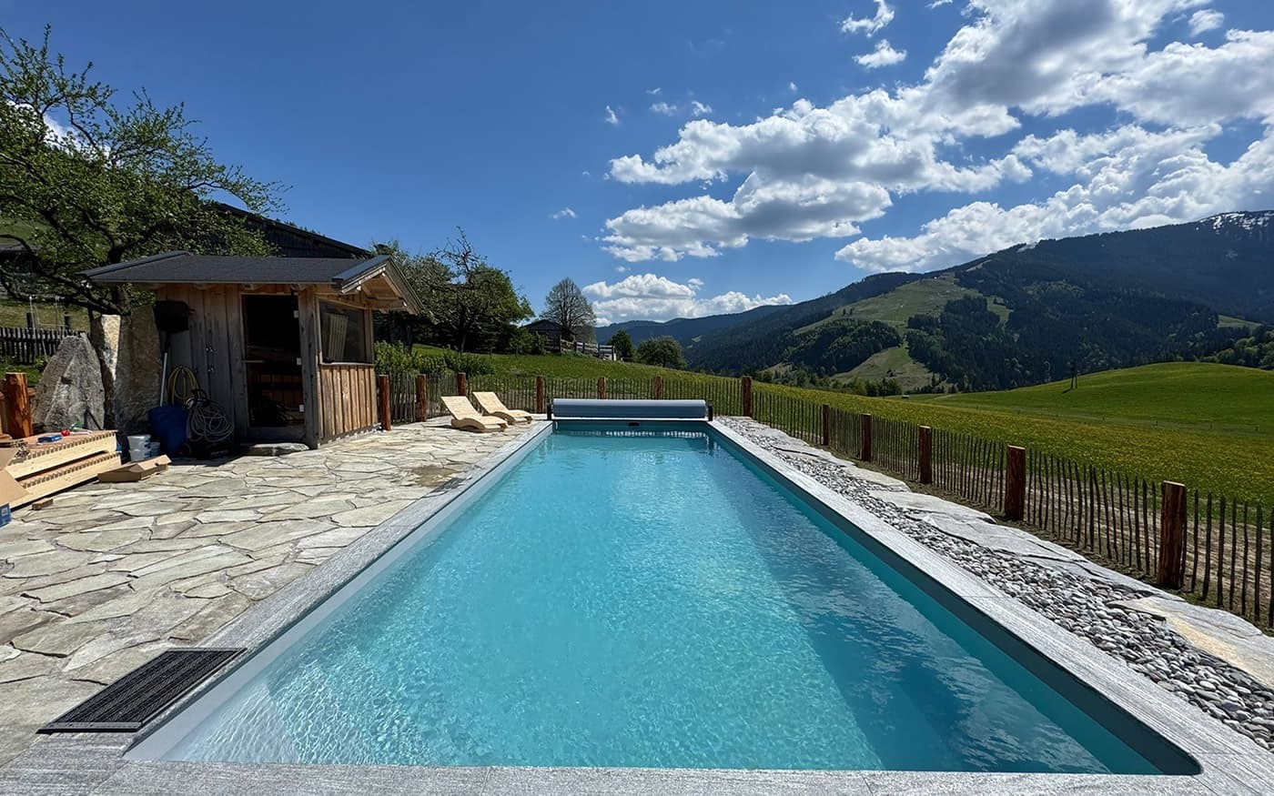 Ein Pool Desjoyaux mit Liegest&uuml;hlen, umgeben von einer malerischen Berglandschaft unter blauem Himmel