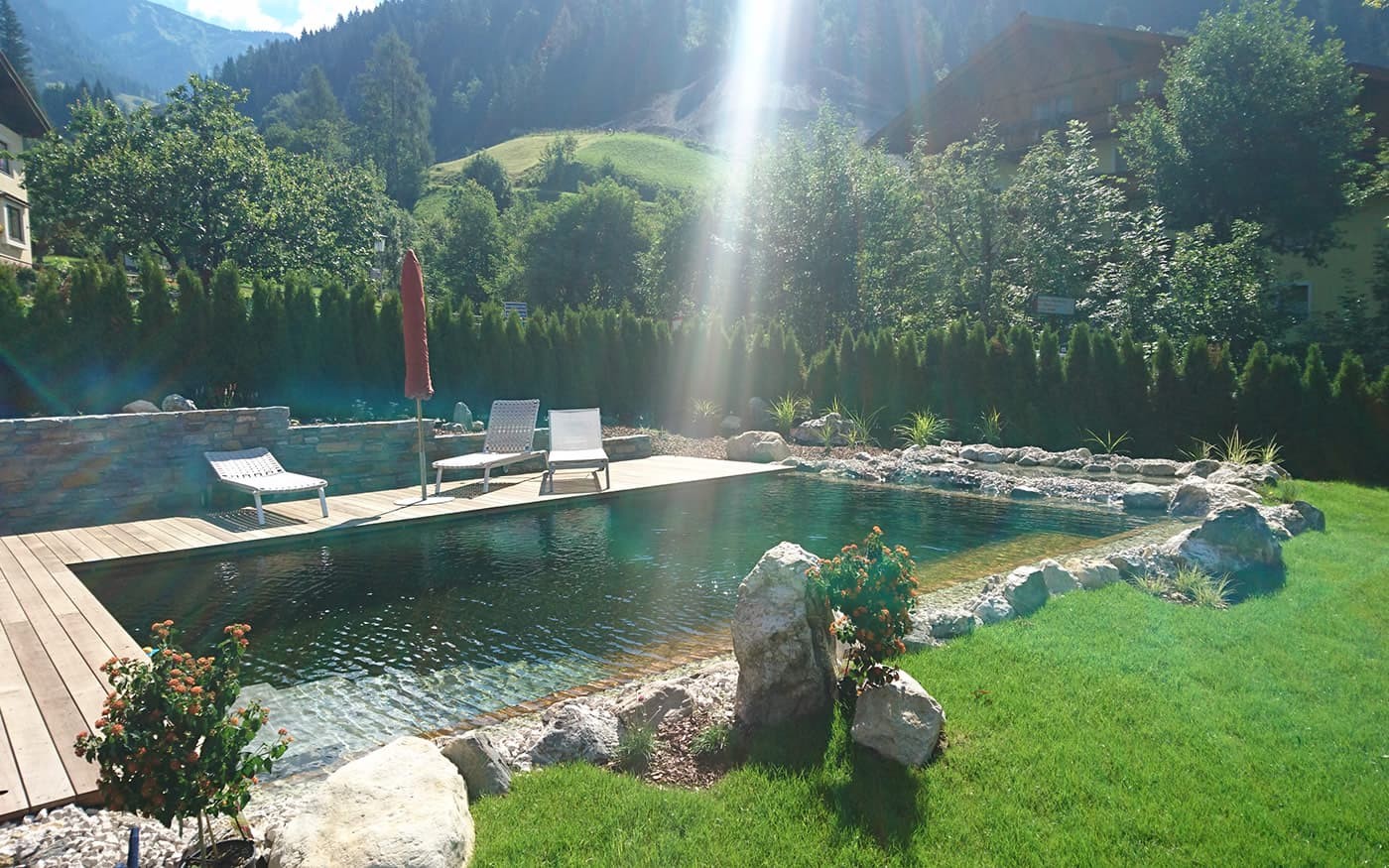 Ein Naturpool mit Liegest&uuml;hlen in einem sonnigen, gr&uuml;nen Garten vor bewaldeten H&uuml;geln
