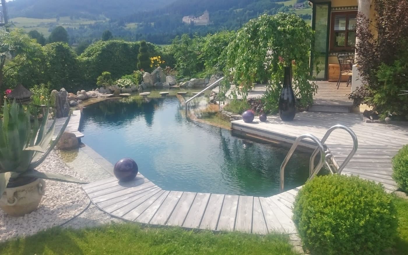 Holzterrasse und Naturteich in einem &uuml;ppigen Garten mit Bergblick im Hintergrund