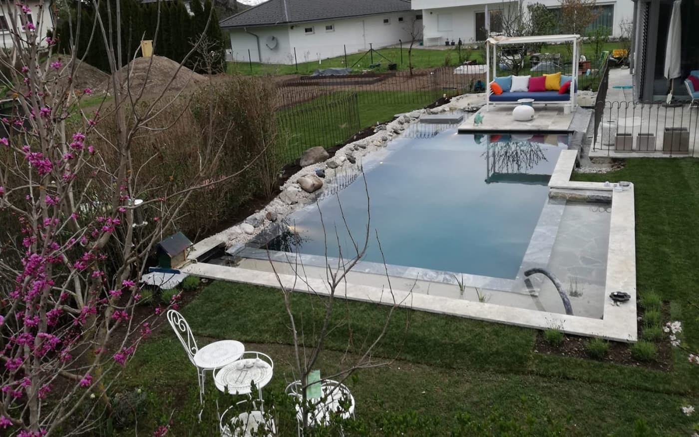 Moderner Garten mit Pool, wei&szlig;en M&ouml;beln und bunten Kissen, umgeben von B&auml;umen und Rasen
