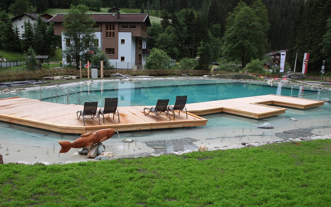 Holzterrasse mit Liegest&uuml;hlen an einem Teich, umgeben von B&auml;umen und Wiese
